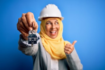 Middle age architect woman wearing muslim hijab and security helmet holding home keys pointing and showing with thumb up to the side with happy face smiling