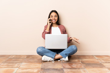 Young mixed race woman with a laptop sitting on the floor keeping a conversation with the mobile phone with someone