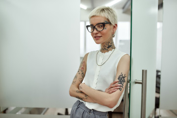I love my job. Young, happy and cute blonde tattooed businesswoman leaning on the glass door, keeping arms crossed and looking at camera with smile while standing in modern office.