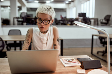 Focused at work. Stylish and beautiful tattooed businesswoman in eyeglasses working with laptop while sitting in the modern office