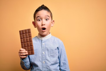 Young little boy kid eating sweet chocolate bar for dessert over isolated yellow background scared in shock with a surprise face, afraid and excited with fear expression