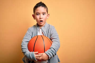 Young little boy kid playing with basketball game ball over isolated yellow background scared in shock with a surprise face, afraid and excited with fear expression