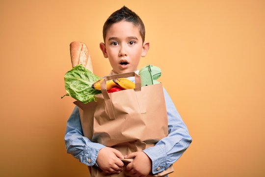 Adorable toddler holding paper bag with food standing over isolated yellow background