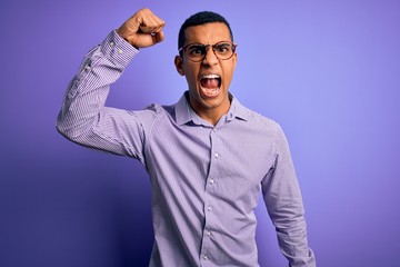 Handsome african american man wearing striped shirt and glasses over purple background angry and mad raising fist frustrated and furious while shouting with anger. Rage and aggressive concept.