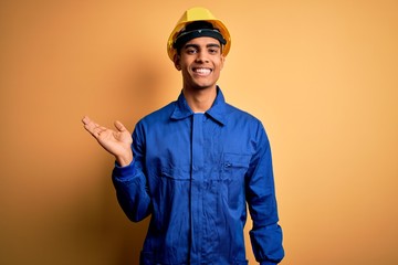 Young handsome african american worker man wearing blue uniform and security helmet smiling cheerful presenting and pointing with palm of hand looking at the camera.