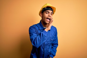 Young handsome african american worker man wearing blue uniform and security helmet hand on mouth telling secret rumor, whispering malicious talk conversation