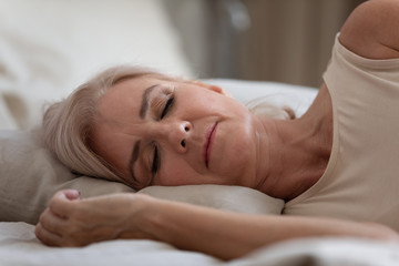 Fototapeta na wymiar Side view serene older woman sleeping on comfortable pillow in bedroom. Head shot close up peaceful middle aged female retiree enjoying sweet dreams at home, resting alone at night or weekend morning.