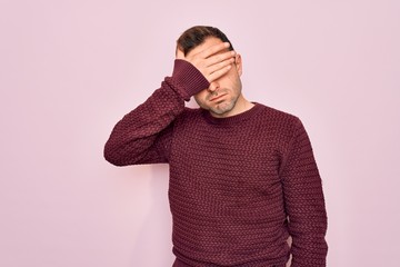 Young handsome man with blue eyes wearing casual sweater standing over pink background covering eyes with hand, looking serious and sad. Sightless, hiding and rejection concept