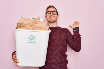 Young handsome man with blue eyes recycling holding wastebasket with cardboard celebrating victory...