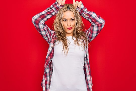 Young beautiful blonde woman wearing casual shirt standing over isolated red background doing funny gesture with finger over head as bull horns
