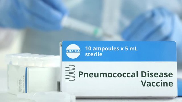 Box with pneumococcal disease vaccine on the table against blurred lab assistant. Fictional phaceutical logo