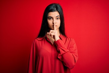 Young beautiful brunette woman wearing casual shirt standing over red background asking to be quiet with finger on lips. Silence and secret concept.