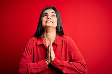 Young beautiful brunette woman wearing casual shirt standing over red background begging and praying with hands together with hope expression on face very emotional and worried. Begging.