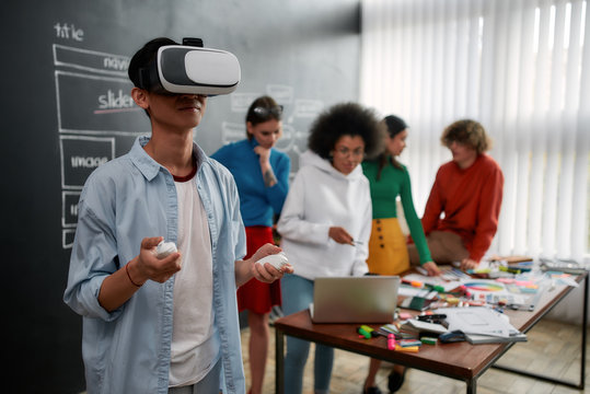 Using modern technologies. Young asian man in casual clothes wearing virtual reality glasses while his colleagues discussing new project behind him