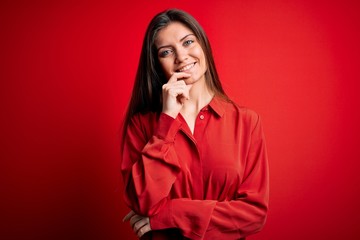 Young beautiful woman with blue eyes wearing casual shirt standing over red background looking confident at the camera with smile with crossed arms and hand raised on chin. Thinking positive.