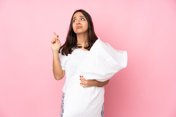 Young woman in pajamas isolated on pink background with fingers crossing and wishing the best
