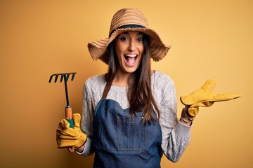 Young beautiful farmer woman wearing apron and hat using rake over yellow background very happy and...
