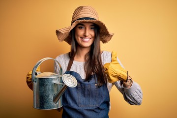 Young beautiful brunette farmer woman wearing apron and hat holding watering can happy with big...
