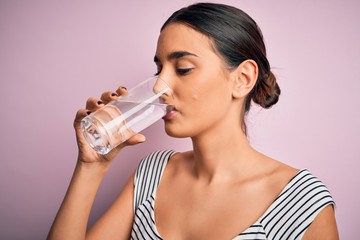 Young beautiful brunette woman drinking glass of healthy water to refreshment standing over isolated pink background