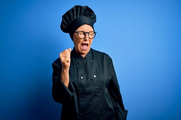 Senior beautiful grey-haired chef woman wearing cooker uniform and hat over blue background angry and mad raising fist frustrated and furious while shouting with anger. Rage and aggressive concept.