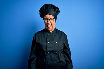 Senior beautiful grey-haired chef woman wearing cooker uniform and hat over blue background sticking tongue out happy with funny expression. Emotion concept.