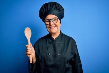Naklejka premium Senior beautiful grey-haired chef woman wearing cooker uniform and hat holding wooden spoon with a happy face standing and smiling with a confident smile showing teeth