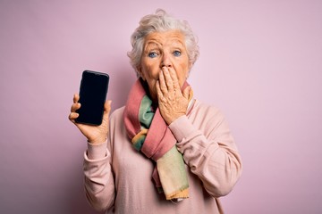 Senior beautiful grey-haired woman holding smartphone showing screen over pink background cover...
