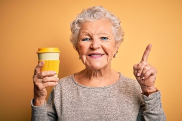 Senior beautiful woman drinking cup of coffee standing over isolated yellow background surprised with an idea or question pointing finger with happy face, number one
