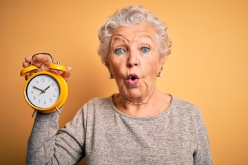 Senior beautiful woman holding alarm clock standing over isolated yellow background scared in shock...