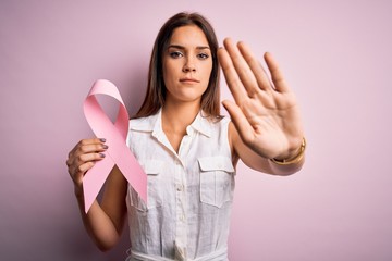 Young beautiful brunette woman holding cancer ribbon symbol over pink background with open hand...