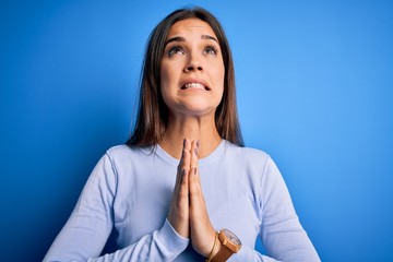 Young beautiful brunette woman wearing casual sweater standing over blue background begging and praying with hands together with hope expression on face very emotional and worried. Begging.