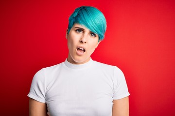 Young beautiful woman with blue fashion hair wearing casual t-shirt over red background In shock face, looking skeptical and sarcastic, surprised with open mouth