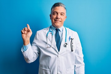 Middle age handsome grey-haired doctor man wearing coat and blue stethoscope with a big smile on face, pointing with hand finger to the side looking at the camera.