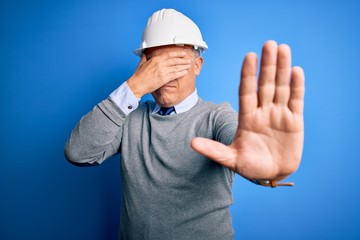 Middle age handsome grey-haired engineer man wearing safety helmet over blue background covering eyes with hands and doing stop gesture with sad and fear expression. Embarrassed and negative concept.