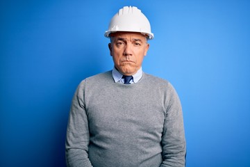 Middle age handsome grey-haired engineer man wearing safety helmet over blue background depressed and worry for distress, crying angry and afraid. Sad expression.