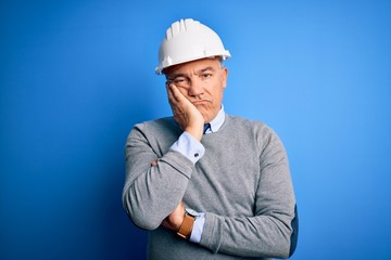 Middle age handsome grey-haired engineer man wearing safety helmet over blue background thinking looking tired and bored with depression problems with crossed arms.