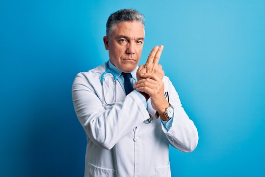 Middle age handsome grey-haired doctor man wearing coat and blue stethoscope Holding symbolic gun with hand gesture, playing killing shooting weapons, angry face