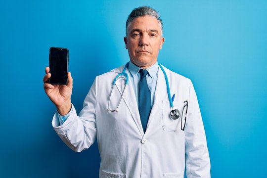 Middle age handsome grey-haired doctor man holding smartphone showing screen with a confident expression on smart face thinking serious