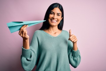 Young beautiful hispanic woman holding paper plane and communication and travel message happy with big smile doing ok sign, thumb up with fingers, excellent sign © Krakenimages.com