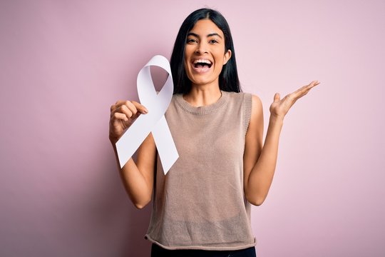 Young beautiful hispanic woman holding white ribbon as stop violence and lung cancer symbol very happy and excited, winner expression celebrating victory screaming with big smile and raised hands