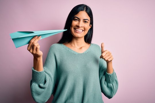 Young beautiful hispanic woman holding paper plane and communication and travel message happy with big smile doing ok sign, thumb up with fingers, excellent sign