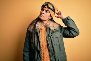 Young beautiful hispanic woman wearing ski glasses and coat for winter weather confuse and...