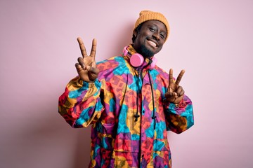 Young handsome african american man wearing colorful coat and cap over pink background smiling looking to the camera showing fingers doing victory sign. Number two.