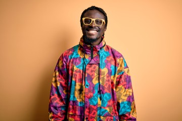 Handsome african american man wearing colorful coat and glasses over yellow background with a happy and cool smile on face. Lucky person.