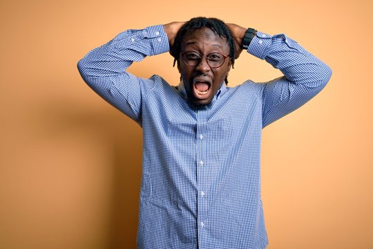 Young handsome african american man wearing shirt and glasses over yellow background Crazy and scared with hands on head, afraid and surprised of shock with open mouth