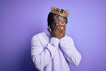 Young african american man wearing golden crown of king over isolated purple background with sad...