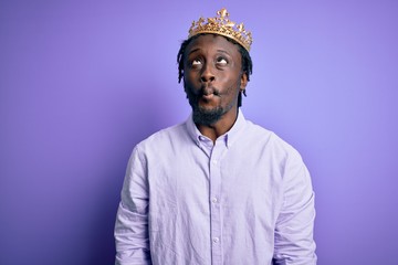 Young african american man wearing golden crown of king over isolated purple background making fish face with lips, crazy and comical gesture. Funny expression.