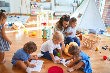 Beautiful teacher and group of toddlers sitting on the floor drawing using paper and pencil around lots of toys at kindergarten - obrazy, fototapety, plakaty