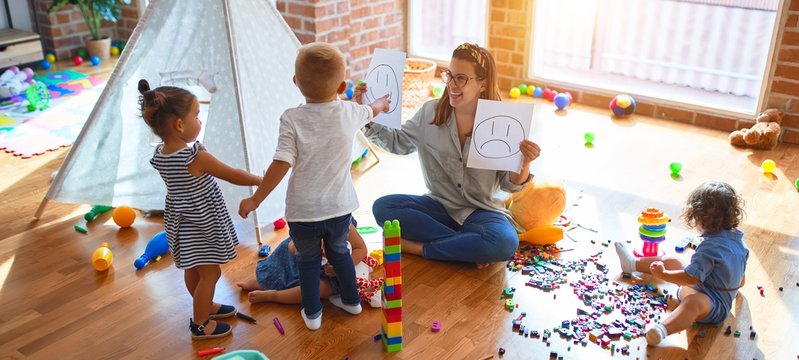 Beautiful psychologist and group of toddlers make therapy using emotions emojis around lots of toys at kindergarten