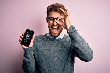 Young man with beard wearing glasses holding broken and craked smartphone with happy face smiling...